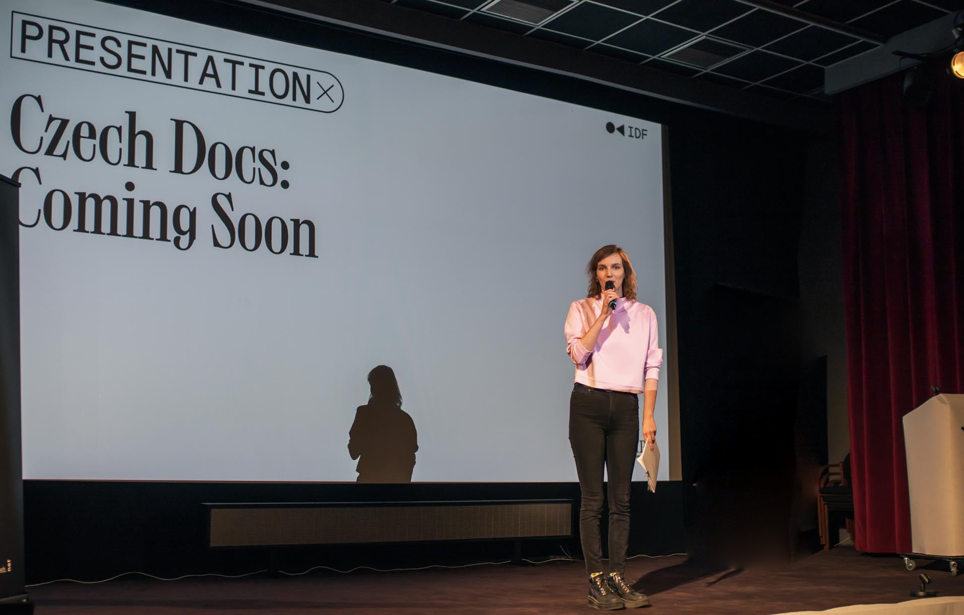Czech Docs Coming Soon Žena stojí na podiu kina a mluví do mikrofonu.