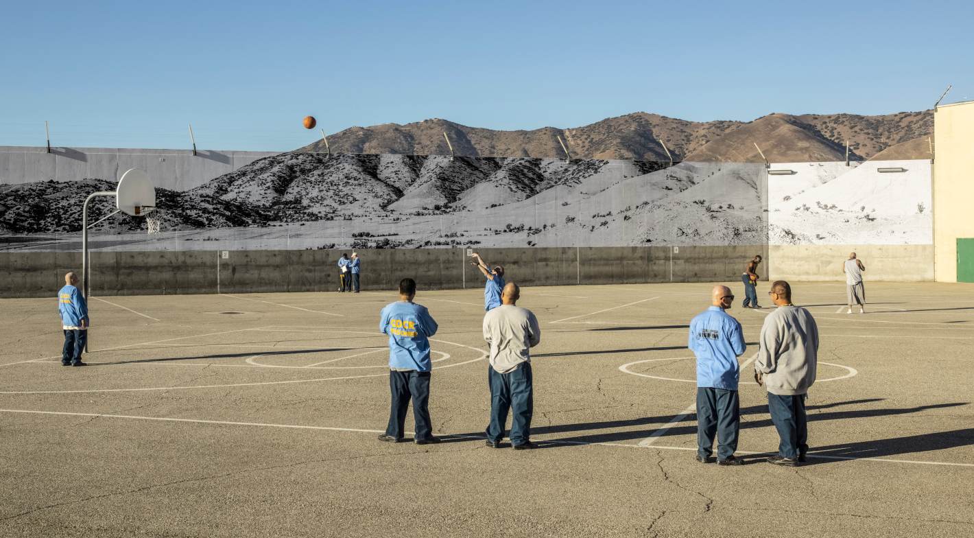 Vězni hrají basketbal na hřišti venku s horami v pozadí. / Inmates playing basketball in a prison yard with mountains in the background.