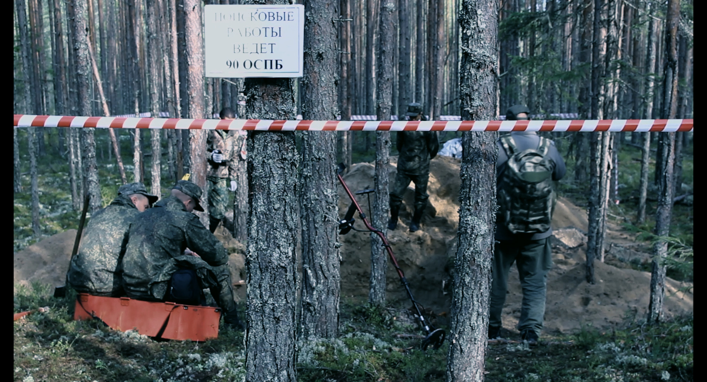 Skupina mužů v maskáčích v lese, před nimi díry, hromady hlíny a detektor kovu.