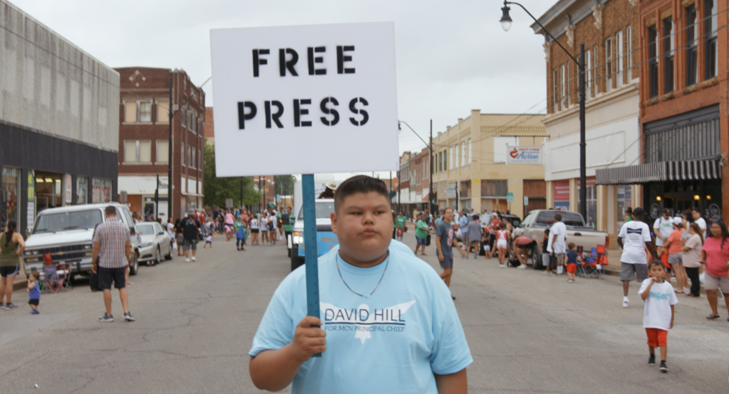 Mladý chlapec stojí uprostřed ulice s transparentem FREE PRESS. Kolem něj jsou  všude na ulici lidé. 