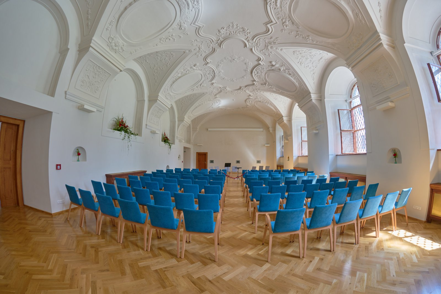Sál refektáře jezuitské koleje s ozdobným štukovým stropem, v sále je několik řad modrých polstrovaných židlí.