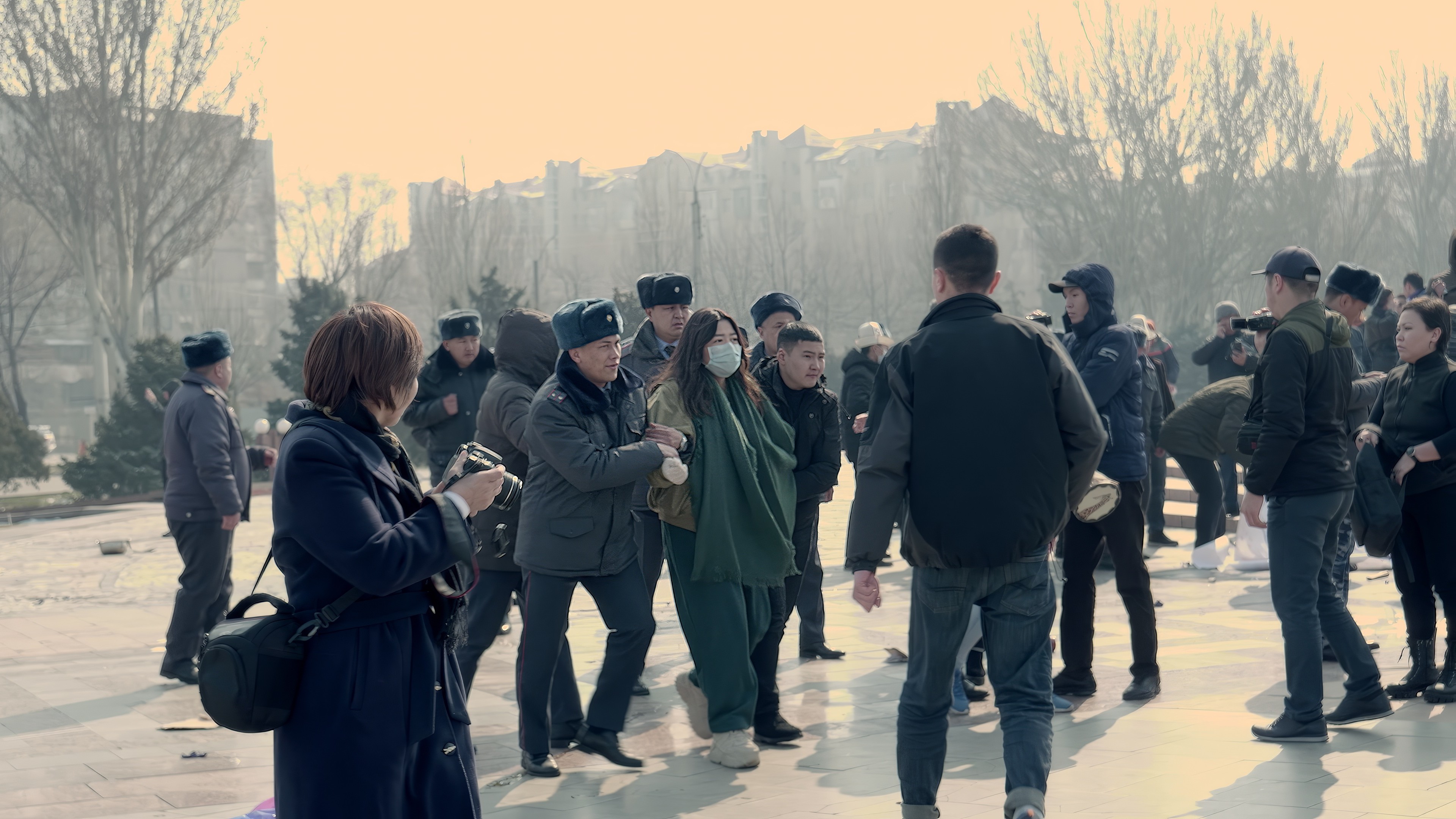 Máte právo vědět Skupina lidí venku na náměstí, někteří fotografují, jiní se procházejí, dva muži vedou ženu v roušce.