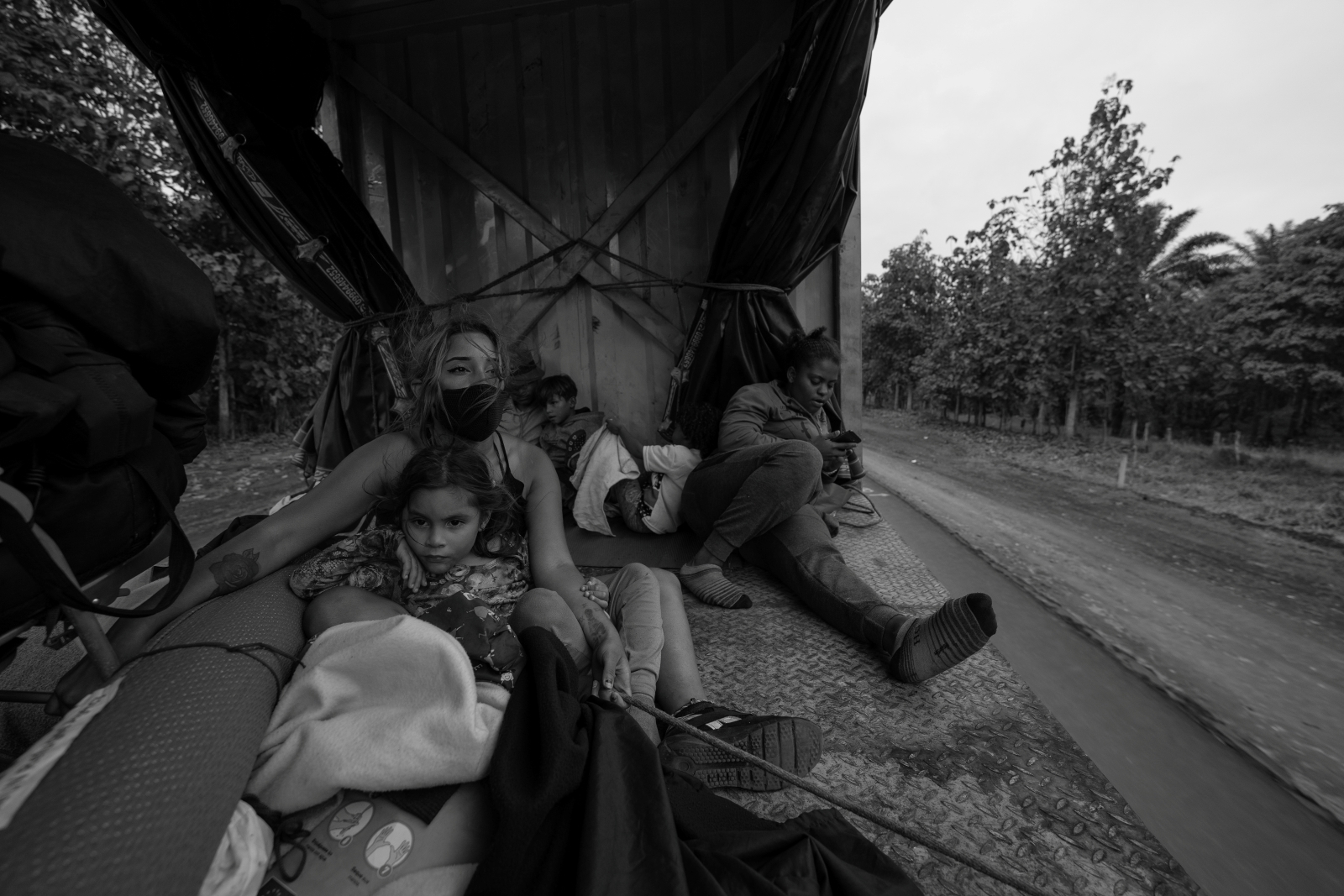 Každý den odvahu Skupina lidí sedí na zadní části nákladního vozu, obklopená zavazadly, na venkovské cestě.