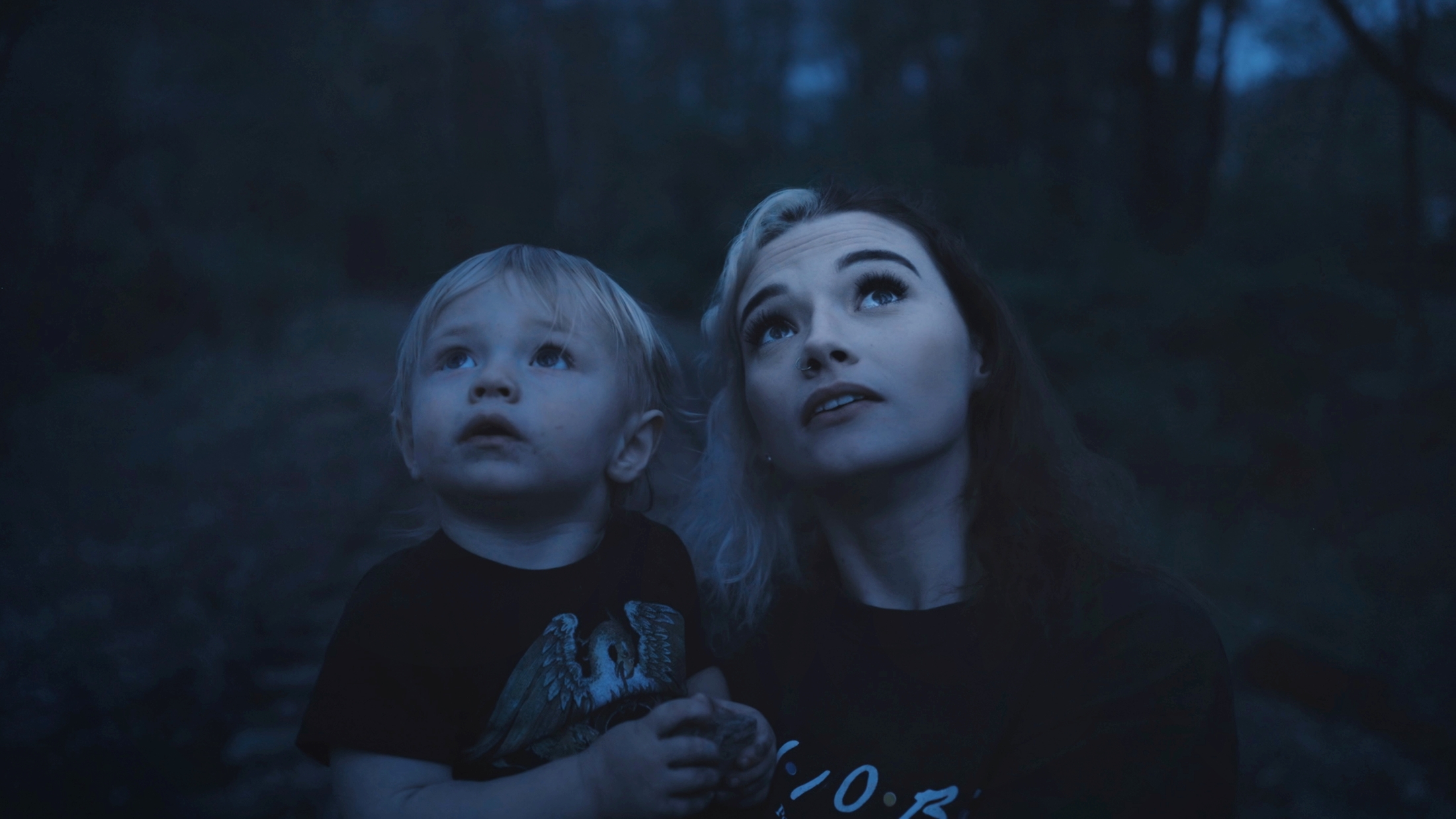 Vize přítomnosti Dospělá osoba a malé dítě v šeru lesa společně vzhlížejí vzhůru, nasvícení jemným modrým světlem.