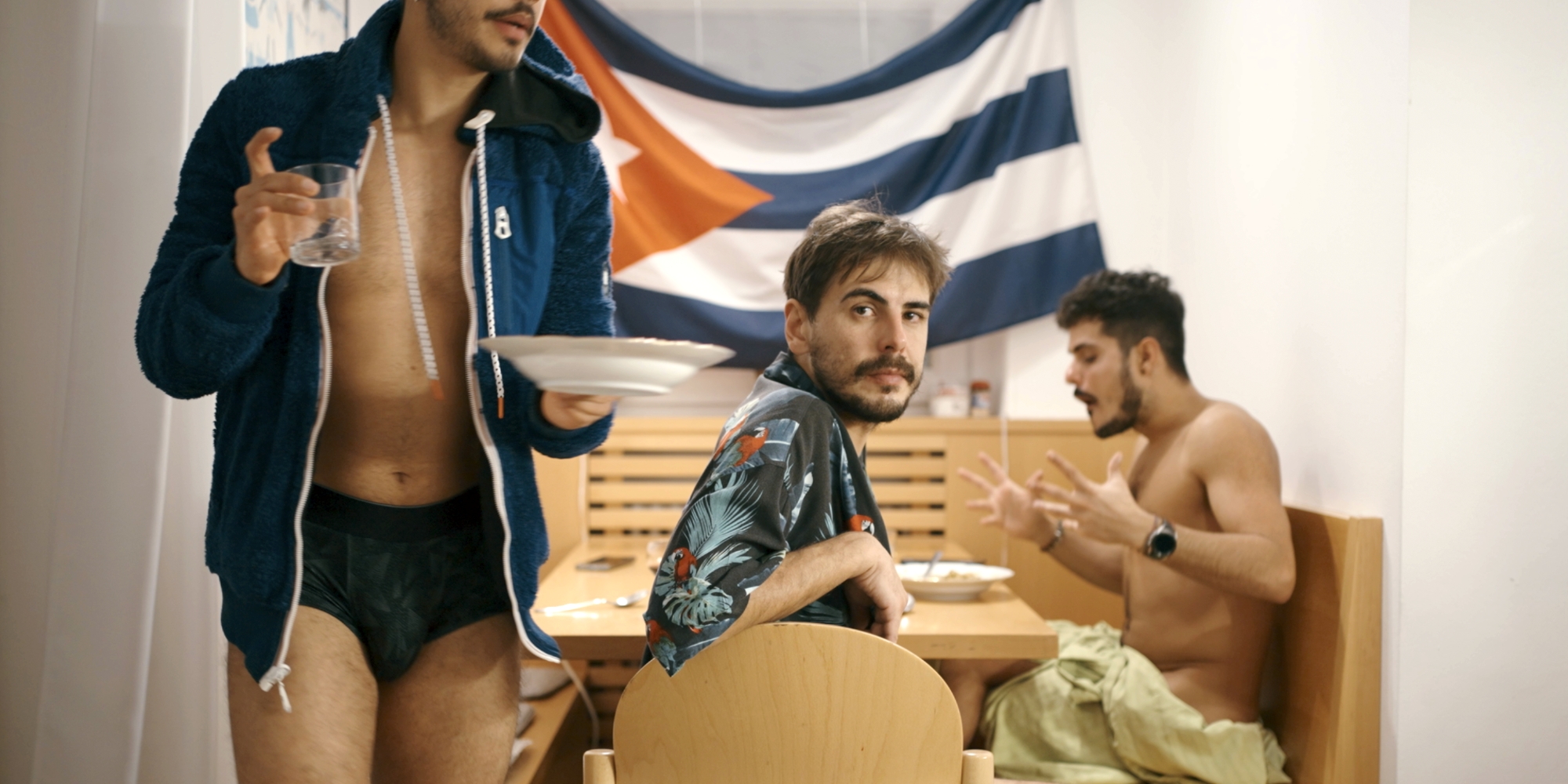 Všechno póly světa Dva muži sedí u stolu v interiéru u jídla, jeden odchází s talířem a sklenicí. V pozadí visí pruhovaná vlajka.