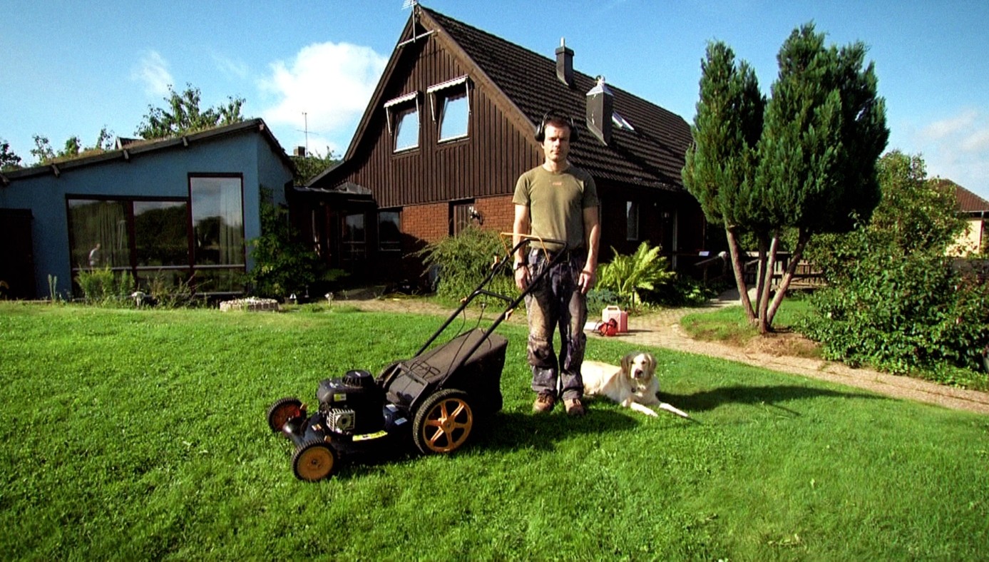 Mýtus Gunnar Muž stojí na zahradě u sekačky na trávu, vedle něj leží pes a v pozadí jsou rodinné domy.