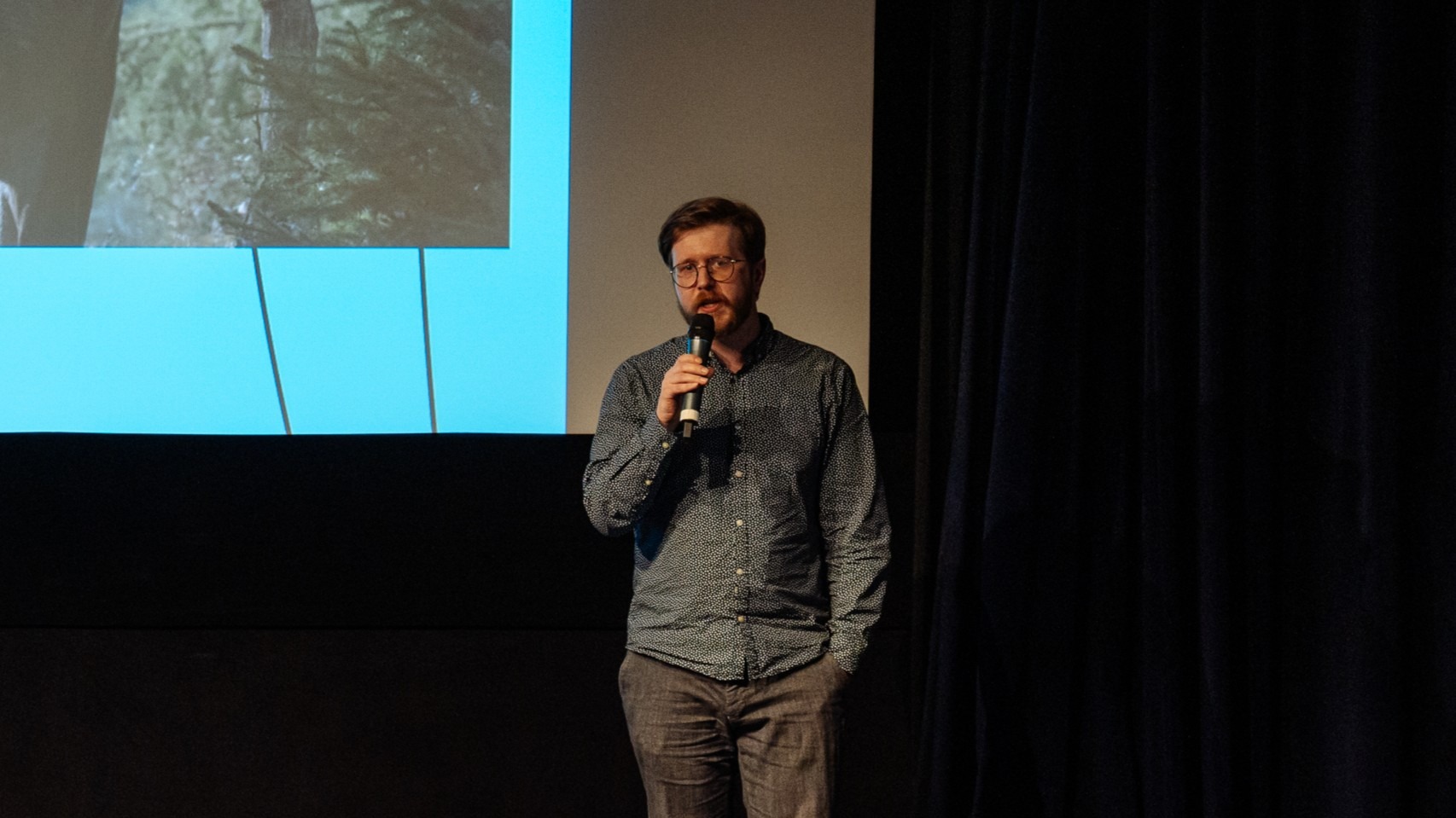 Muž stojící před filmovým plátnem mluví do mikrofonu