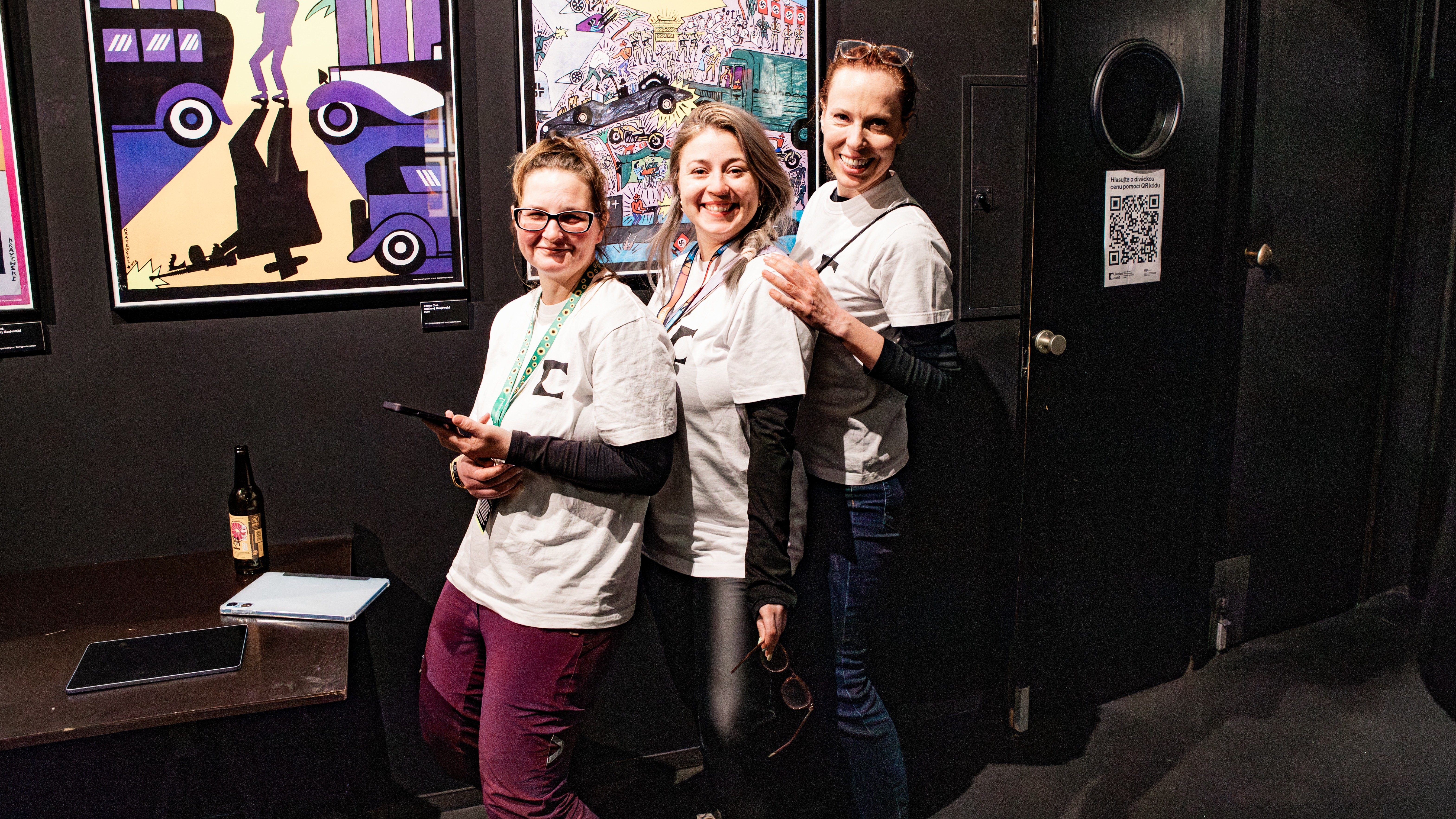 Tři mladé ženy stojí za sebou a se usmívají se. Na sobě mají trička s logem filmového festivalu Jeden svět a na krku pověšené cedulky Pořadatel.