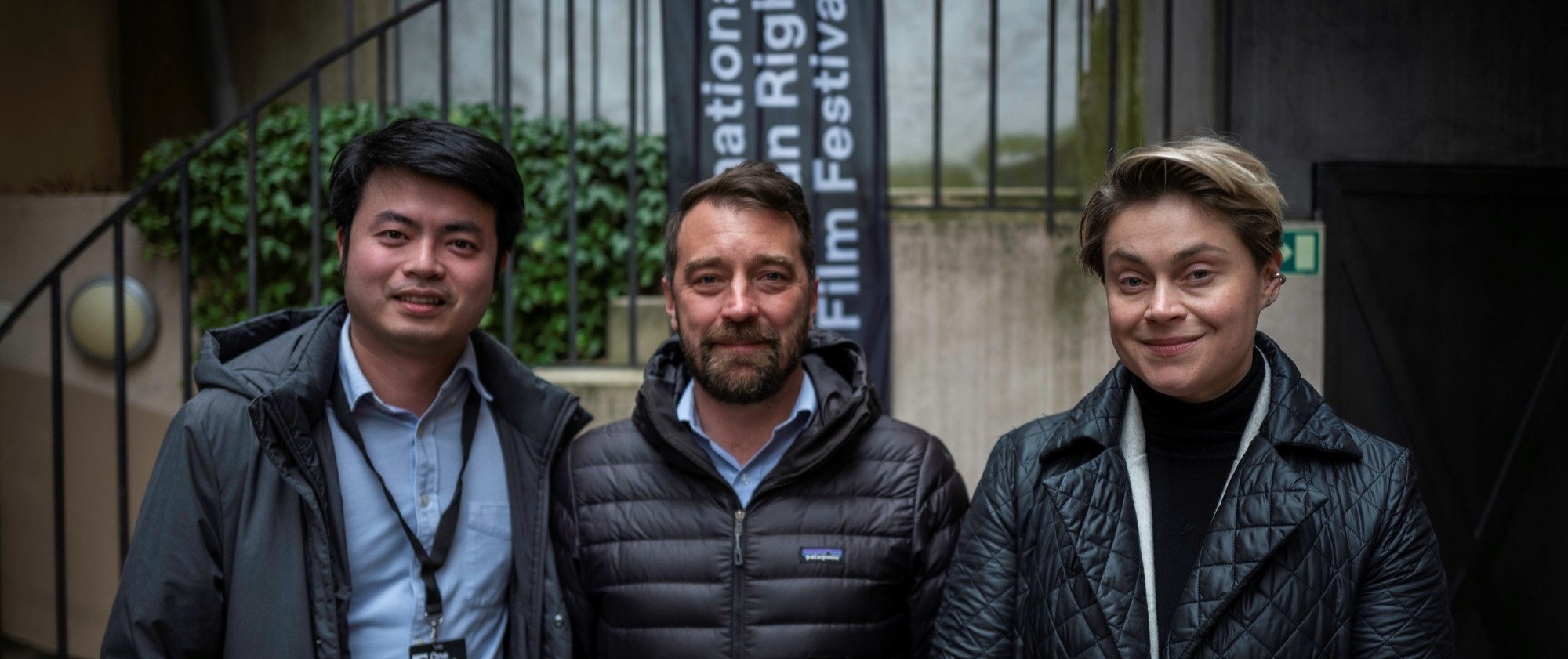 Tři osoby stojí vedle sebe. Za nimi je schodiště s kovovým zábradlím.