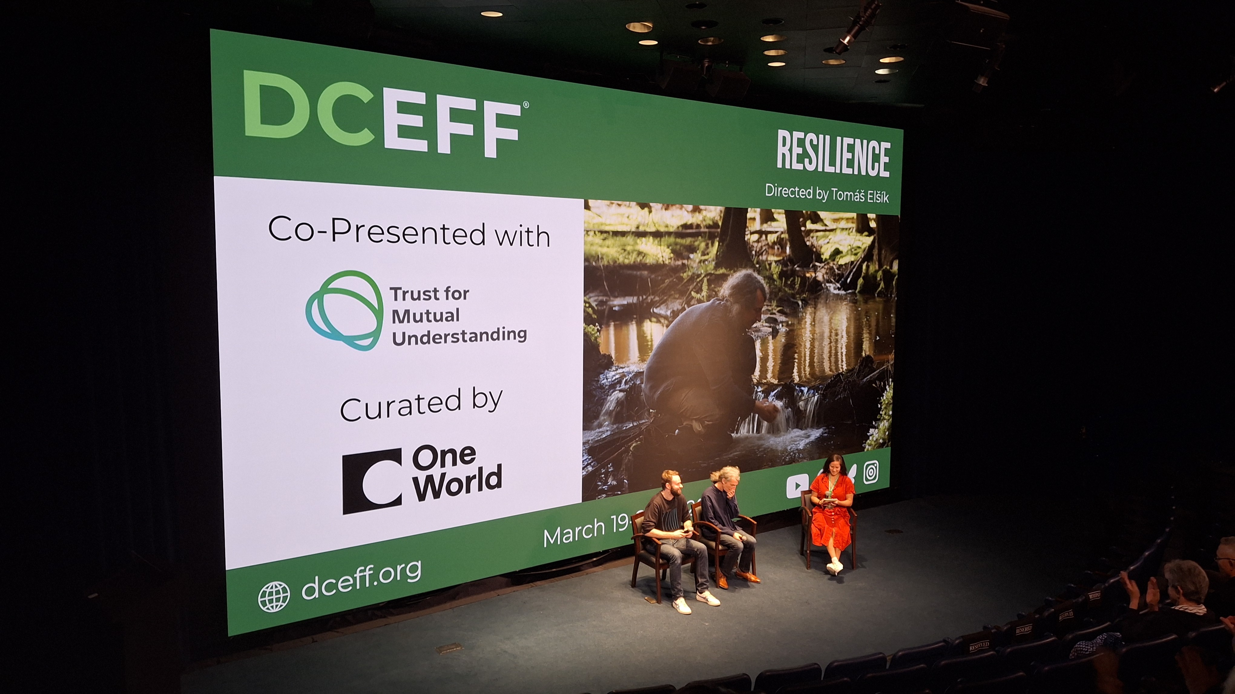 Dva muži a žena sedí ve velkém sále před projekčním plátnem s názvem festivalu DCEEF a filmu Resilience.