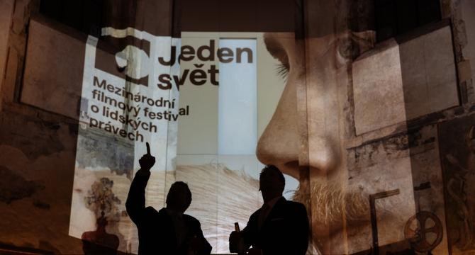 Jeden svět jako pojistka demokracie Dvě postavy stojí ve tmě a kontrastují s projekcí festivalového vizuálu.