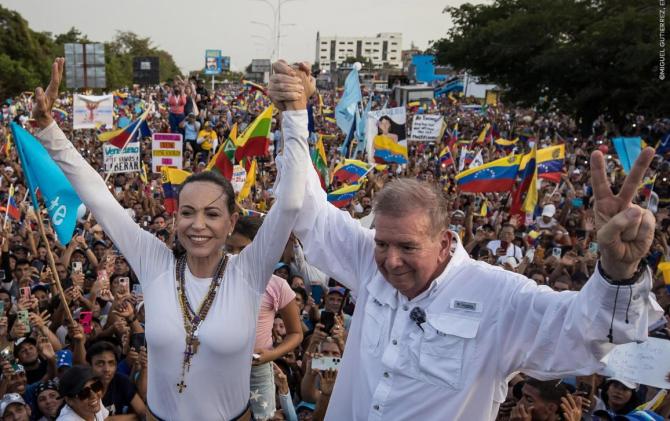 María Corina Machadová a Edmundo González Urrutia získali Sacharovovu cenu za rok 2024 Žena a muž stojí s rukama zvednutýma nad hlavou ve vítězném gestu, za nimi je dav lidí s vlajkami a transparenty.