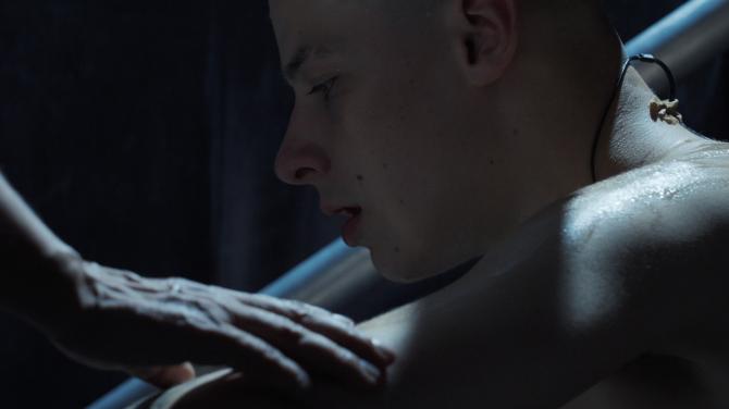 Mladý muž sedí ve tmavém prostředí a někdo mu pokládá ruku na rameno.