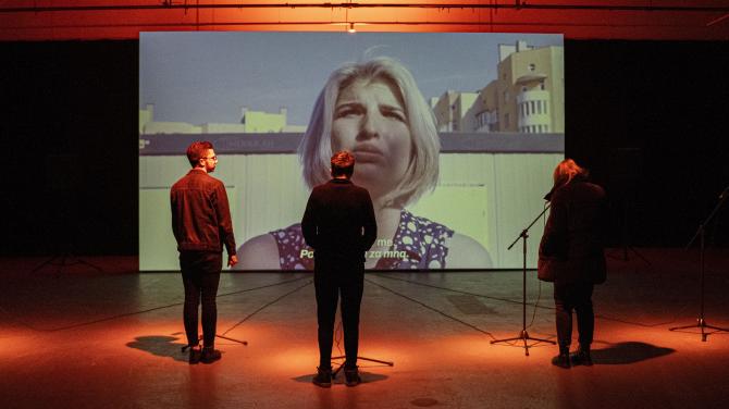 Vidět nestačí. Unikátní projekt Repeat After Me reprodukuje zvukové vjemy války V tmavém sále stojí tři osoby před velkou projekcí ženy s blond vlasy; před každou osobou je mikrofon a prostor je nasvícen oranžovým světlem.