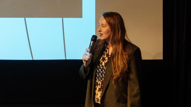 Žena stojící před filmovým plátnem mluví do mikrofonu.