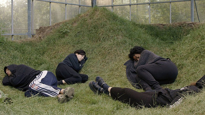 Čtyři lidé leží schoulení na trávě uvnitř proskleného skleníku. Jsou oblečeni převážně v tmavém oblečení a působí, jako by odpočívali nebo spali, zatímco kolem nich je vysoká tráva a skrz skleněné stěny prosvítá světlo z okolních stromů.