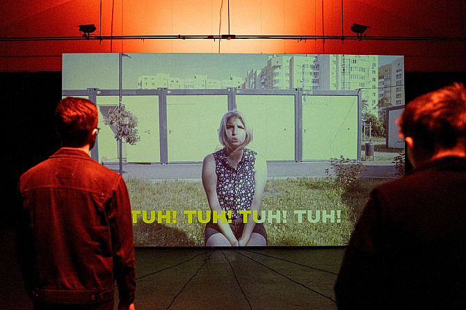 „Dva návštěvníci stojí v temné galerii a sledují velkou projekci videa. Na plátně sedí žena na trávě před panelovými domy a vyslovuje slovo ‚TUH!‘, které se opakuje jako titulky na obrazovce.