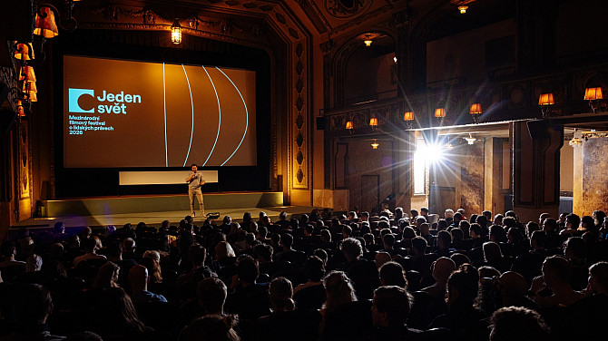 Moderátor stojí na pódiu v historickém kině a hovoří k plnému sálu diváků. Na velkém plátně za ním je promítán vizuál festivalu Jeden svět – Mezinárodní filmový festival o lidských právech 2026.