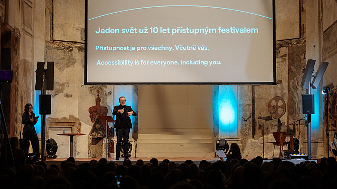 Na pódiu v historickém interiéru stojí muž, který tlumočí do znakového jazyka, vedle něj žena s poznámkami. Za nimi je velké projekční plátno s nápisem o festivalu Jeden svět a sdělením o přístupnosti pro všechny. Před pódiem sedí publikum sledující program.