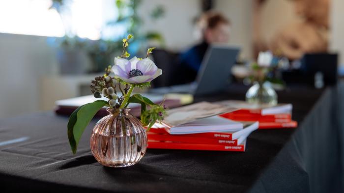 Těšíme se na setkání Na stole s černým ubrusem stojí váza s kyticí a štos katalogů.