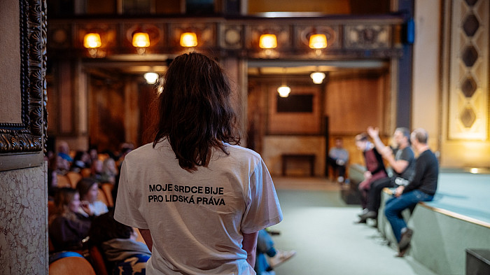 Osoba s delšími vlasy stojí zády k fotoaparátu uprostřed sálu, na sobě má bílé tričko s nápisem „Moje srdce bije pro lidská práva“. Před ní je pódium, kde sedí několik lidí zapojených do diskuse, naproti nim sedí publikum.