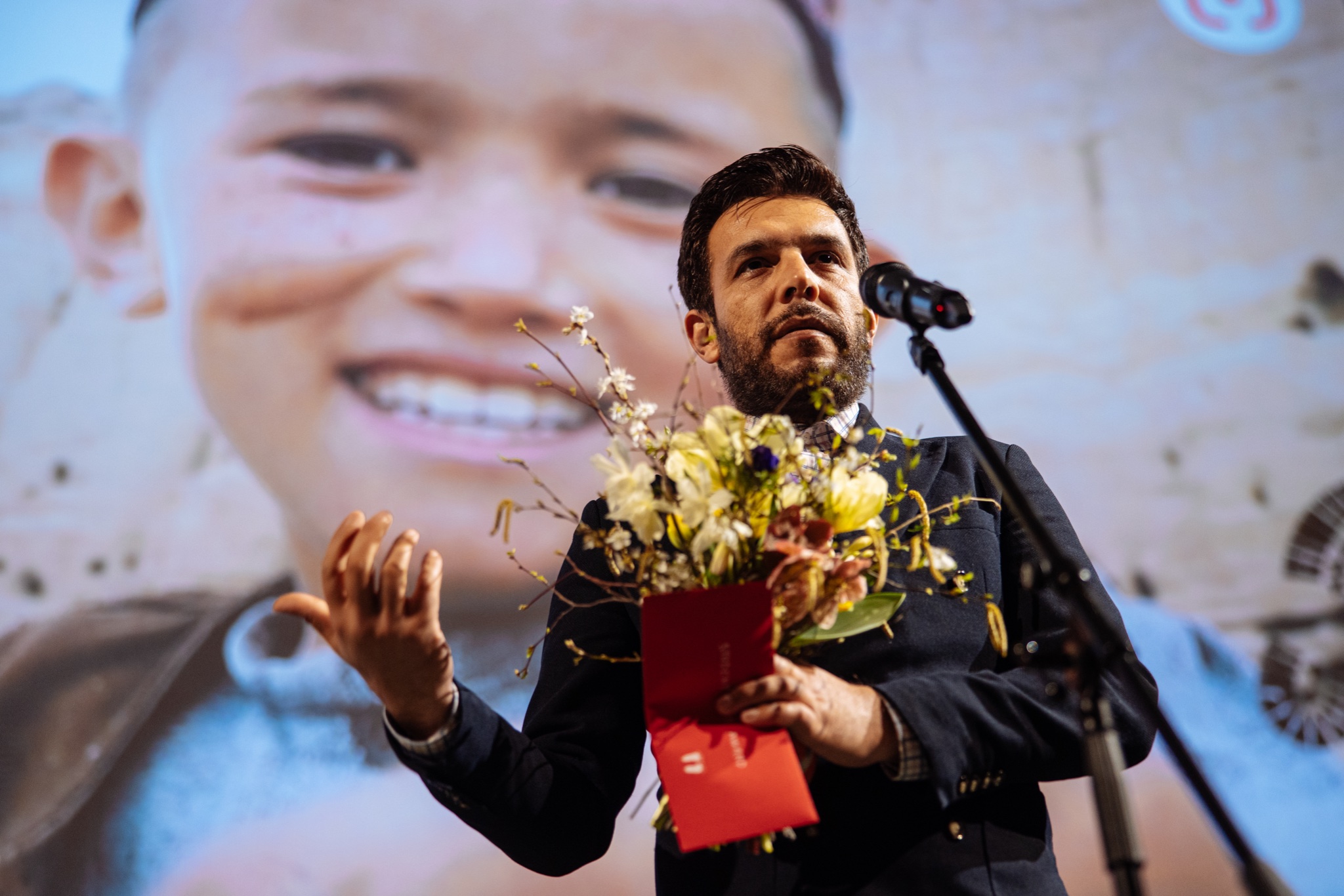 Cenu na podiu přebírá spoluležisér Shoaib Sharifi. Drží květinu a hovoří do mikrofonu.