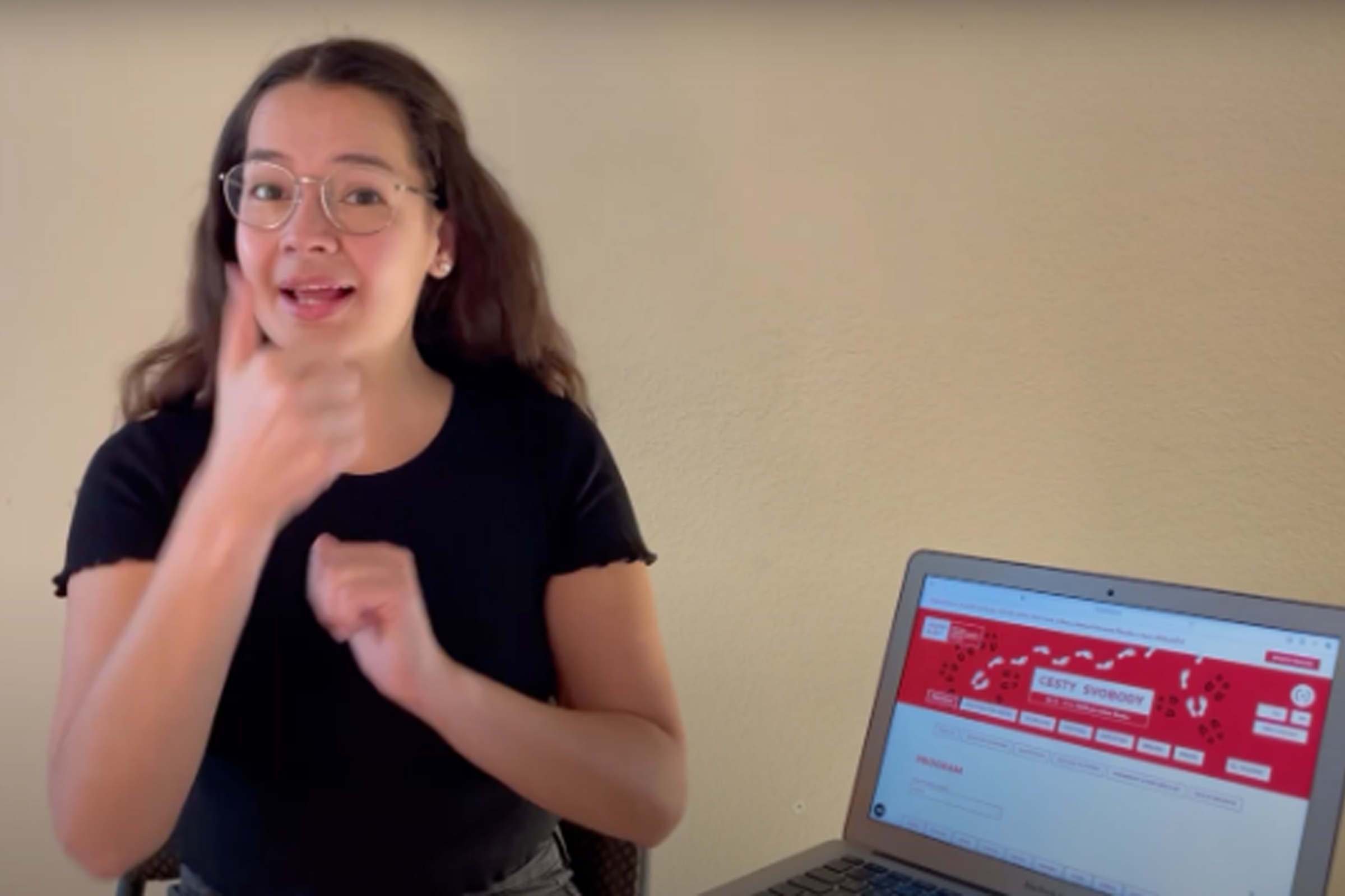 Žena vysvětluje znakovým jazykem, kde lze na webových stránkách nají informace o programu festivalu. Vedle sebe má na stole otevřený počítač s otevřenými stránkami.