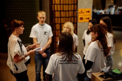 Sedm osob stojí v kruhu ve foyer, jedna z nich drží papír a vysvětluje pokyny.
