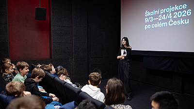 Mladá žena stojí před promítacím plátnem v malém kinosále a mluví ke skupině sedících školních dětí. Na plátně je text „Školní projekce 9/3–24/4 2026 po celém Česku“. Děti sedí v řadách sedaček a poslouchají prezentaci.