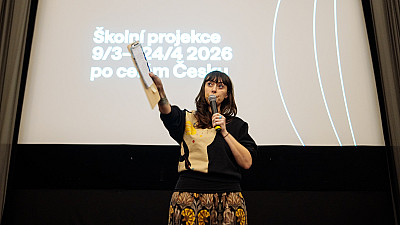 Žena stojí před plátnem, na kterém je napsáno Školní projekce 9/3 - 24/4 2026 po celém Česku, ukazuje a mluví k publiku do mikrofonu.