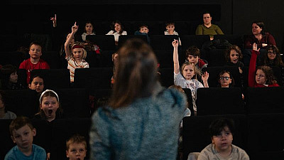 Skupina dětí sedí v kinosále, některé zvedají ruku, zatímco vpředu stojí dospělá osoba zády k nim, pravděpodobně vedoucí nebo lektorka, která s nimi komunikuje.