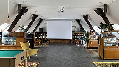 Podkrovní interiér s dřevěnými trámy, uprostřed promítací plátno, po stranách knihovny a vitríny s předměty; vlevo kulečníkový stůl a žluté křeslo.