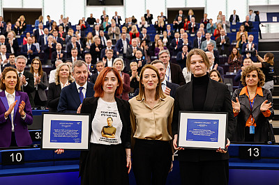 Tři osoby stojí v popředí a drží zarámované dokumenty, zatímco za nimi sedí a stojí zaplněný sál lidí, část publika tleská. Scéna působí jako oficiální událost nebo ceremonie ve velkém konferenčním prostoru.